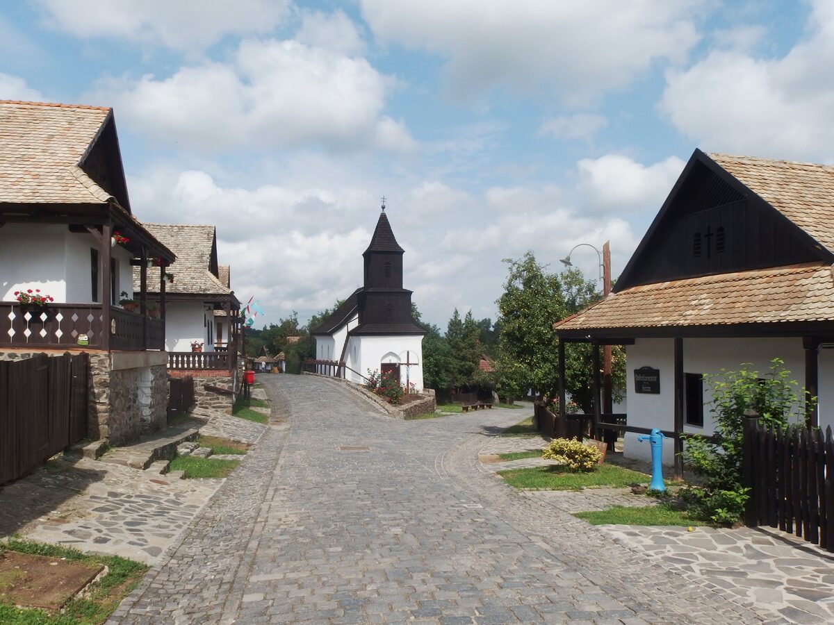 A cobbled street in Hollókő's old village between whitewashed Palóc houses with wooden gables, leading to the small church with its wooden tower