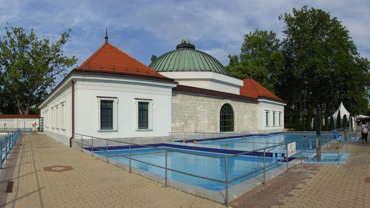 Outdoor pools of the Eger Thermal Bath beside a white bath house with a green copper dome, among tall trees