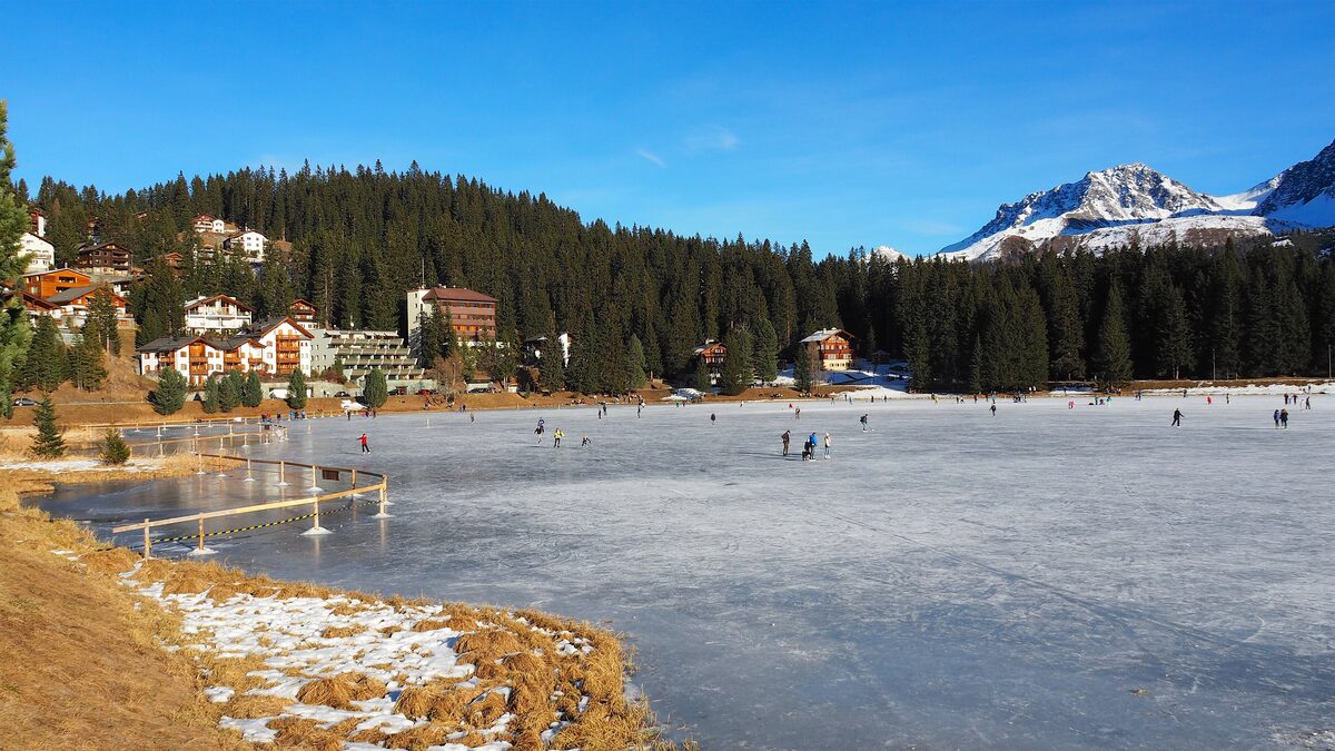 Skaters on the frozen Obersee at Arosa, with chalets and hotels under pine forest and a snowy peak behind