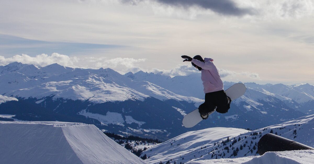 A snowboarder in mid-air over a LAAX park kicker, with snowy peaks behind