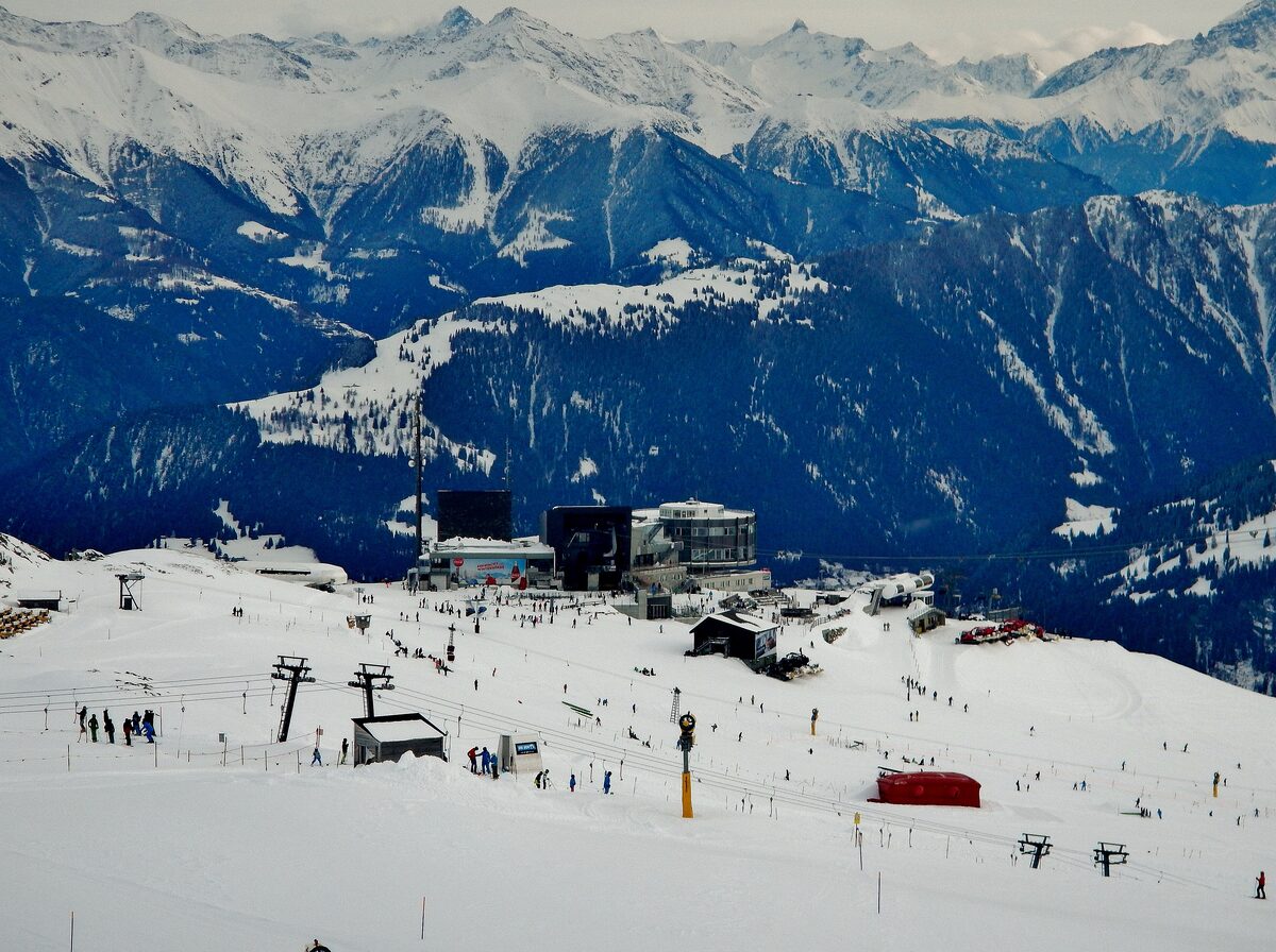 The Crap Sogn Gion summit station in winter, with skiers on the snowy slopes below and the peaks behind