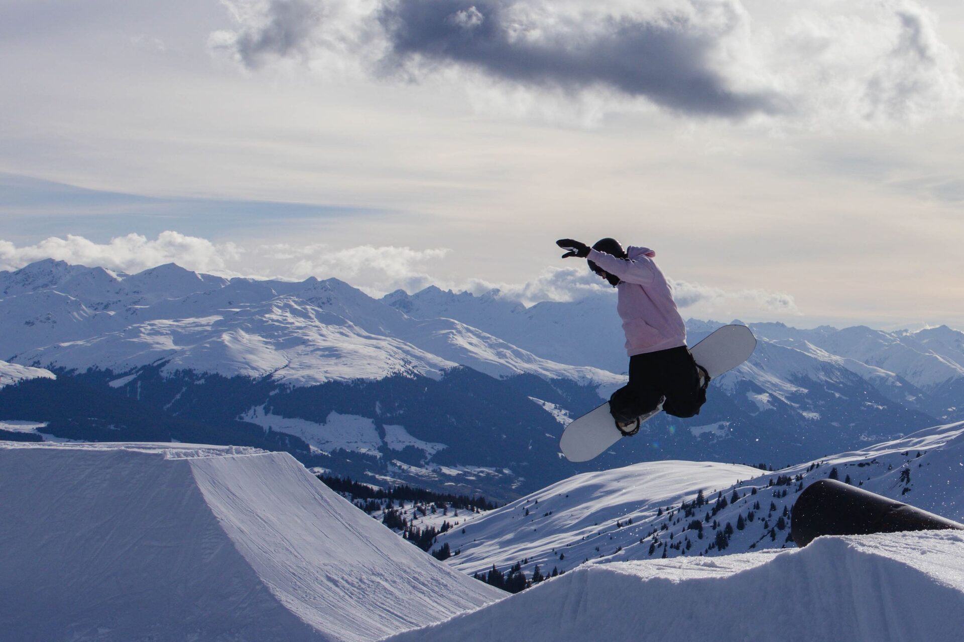 A snowboarder in mid-air over a LAAX park kicker, with snowy peaks behind