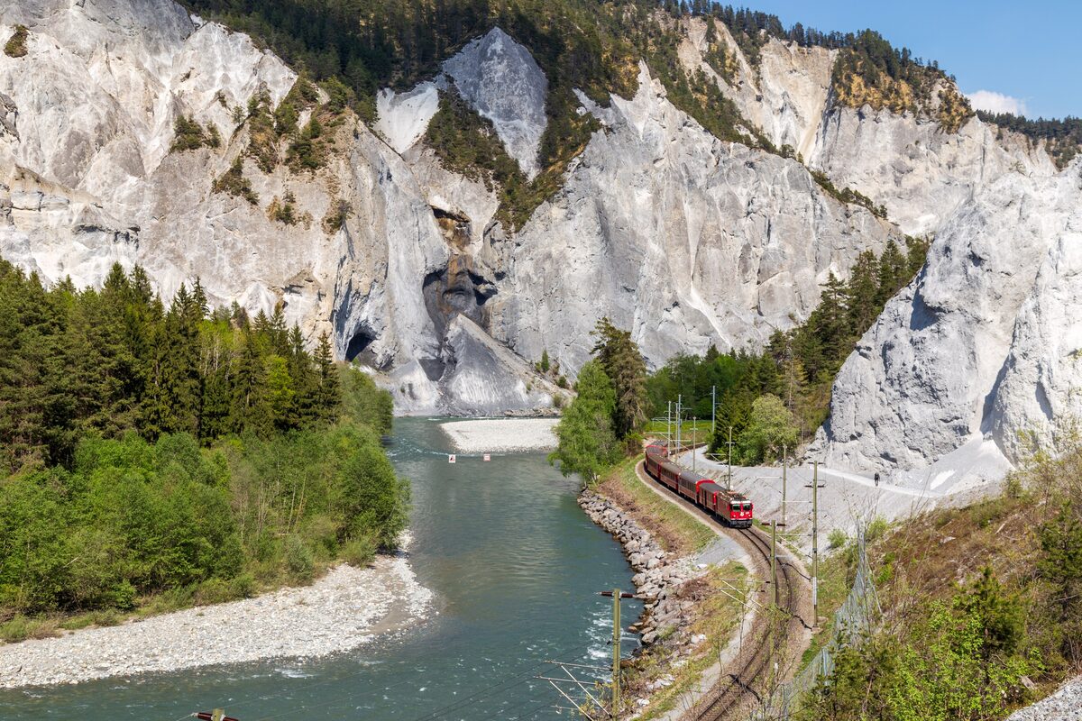 A red Rhaetian Railway train running beside the Rhine between pale cliffs and dark pines