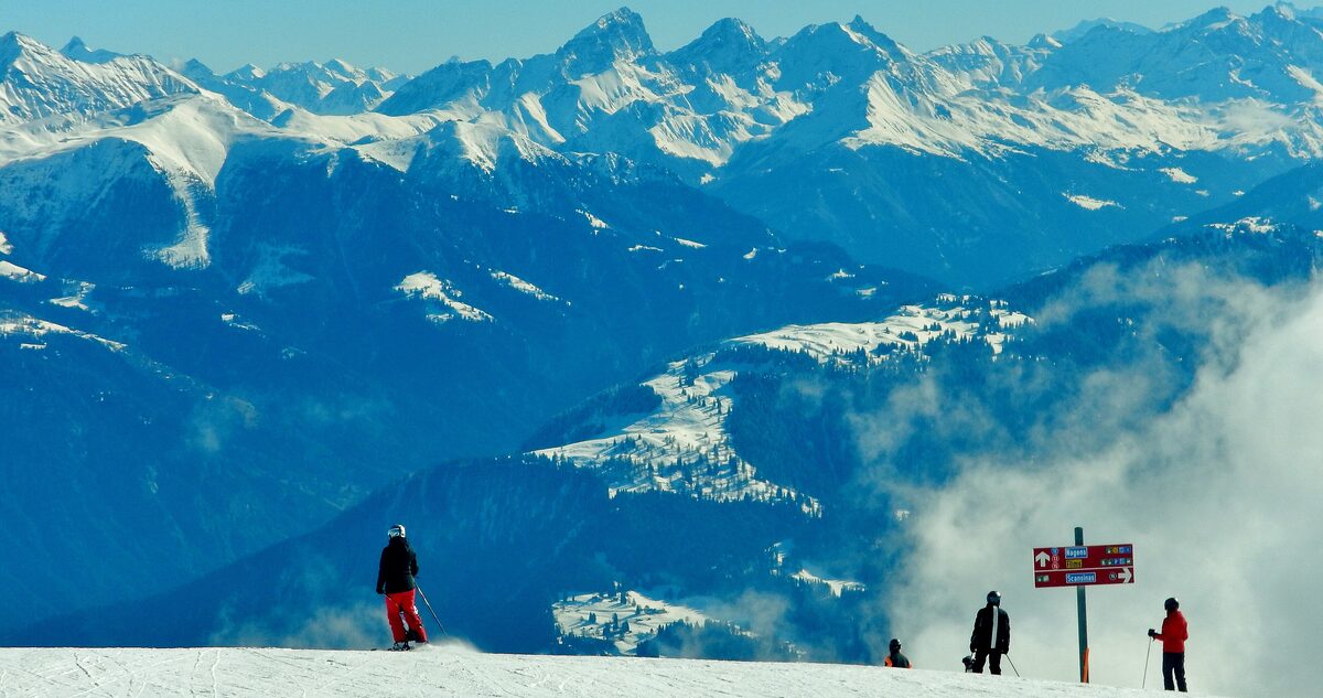 Skiers at a piste marker on a snowy ridge above the clouds, with the Alps across the valley