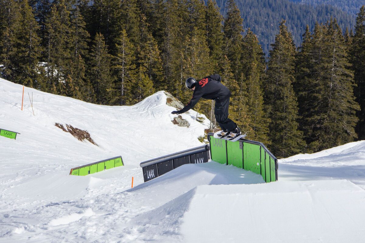 A snowboarder sliding a green rail in the LAAX snowpark, with pine forest above
