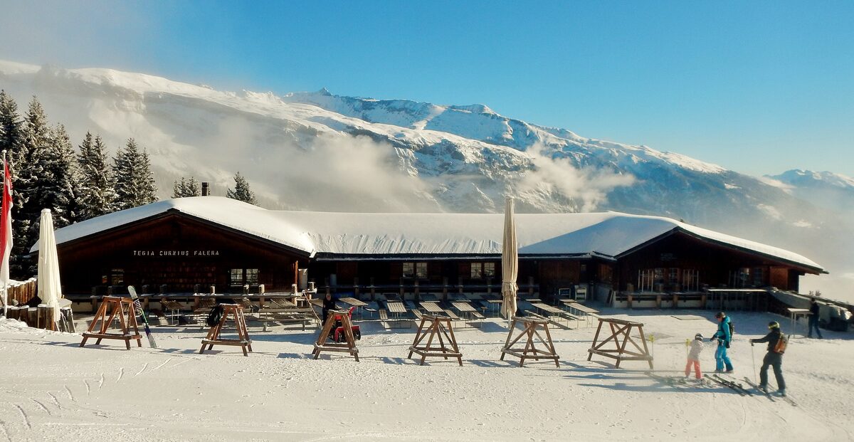 The Tegia Curnius restaurant in snow, with a sunlit terrace, skiers and a mountain range behind