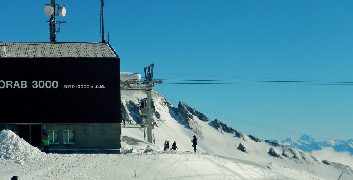 The Vorab 3000 mountain station, a black building in snow under a blue sky, with skiers on the slope