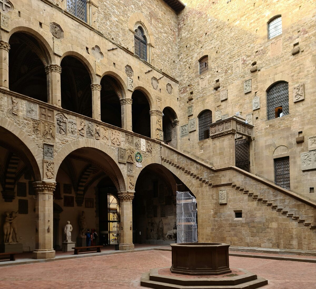 The medieval courtyard of the Bargello, with stone arches, coats of arms on the walls, an open staircase and a well in the middle