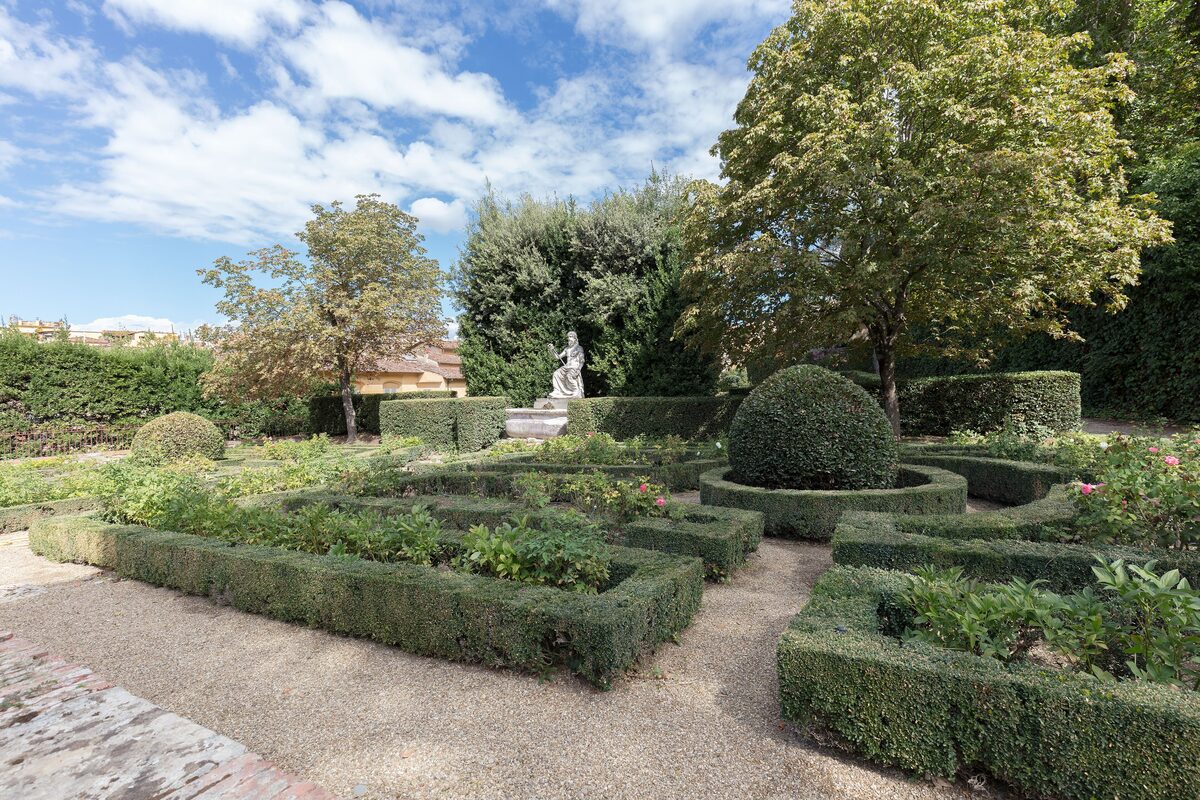 A formal garden in Boboli with low clipped box hedges around flower beds, trees and a statue in the background