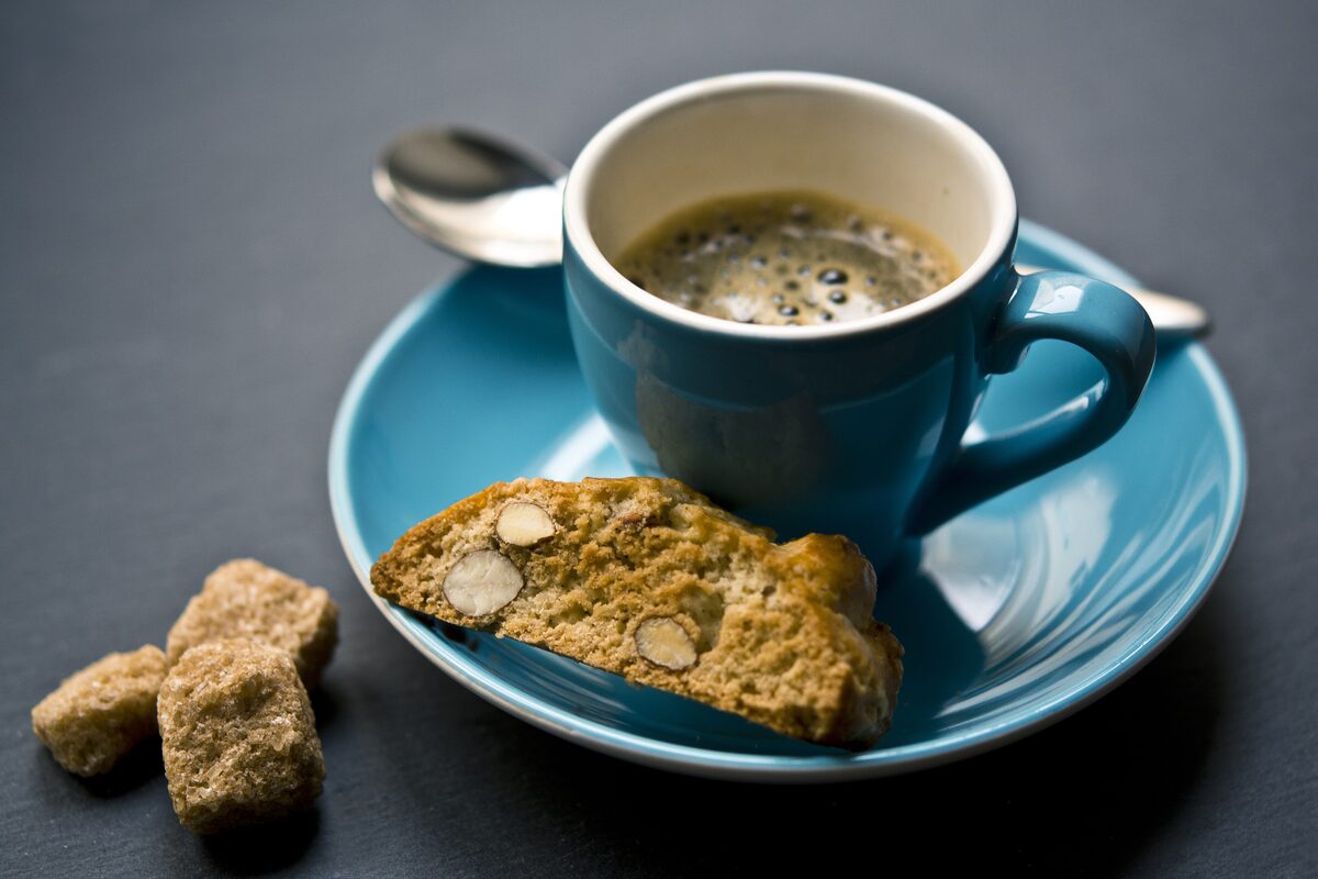 An almond cantuccio biscuit on a blue saucer next to a cup of espresso and two brown sugar cubes
