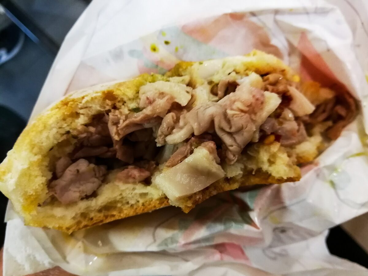 A lampredotto sandwich in a bun, filled with chopped tripe and green sauce, wrapped in paper