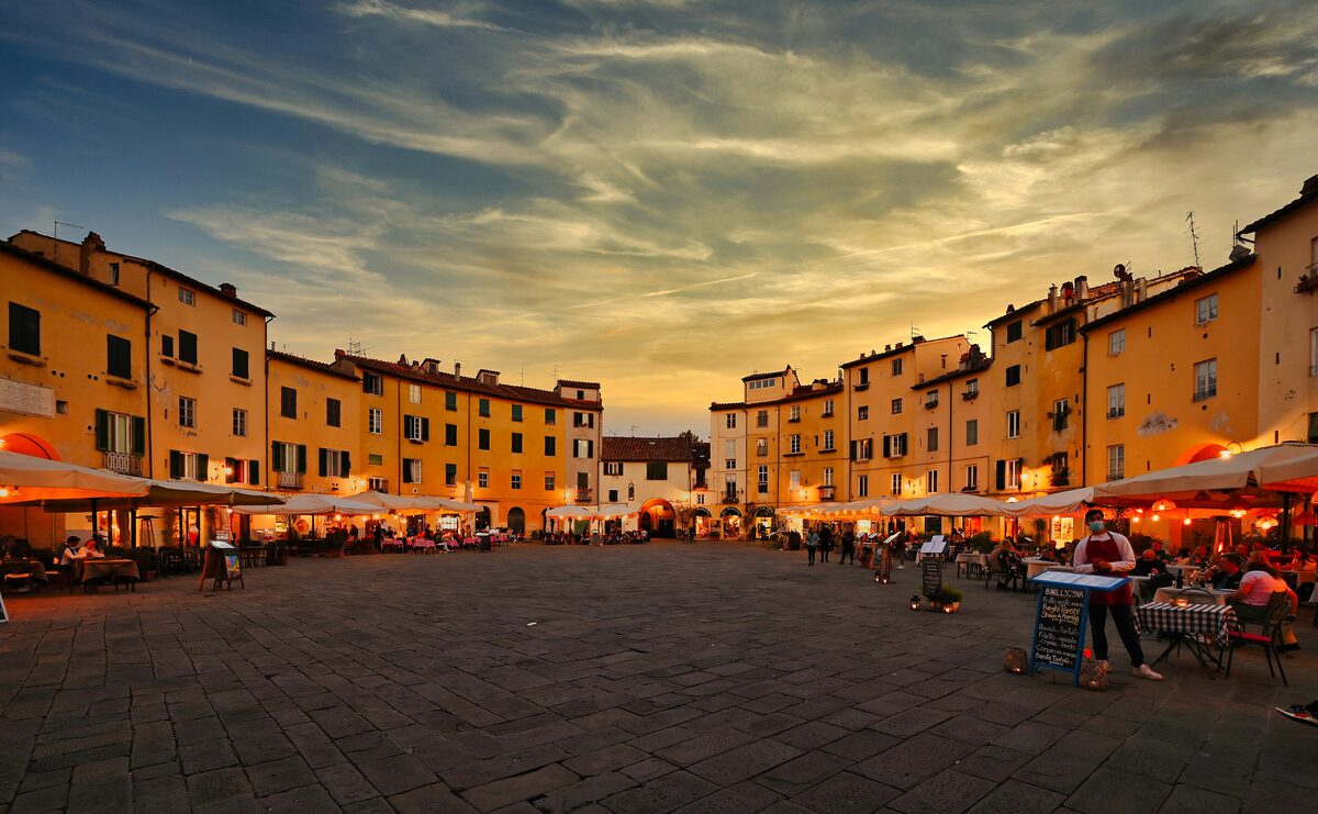 The oval Piazza dell'Anfiteatro in Lucca at sunset, ringed by old houses with restaurant tables in front