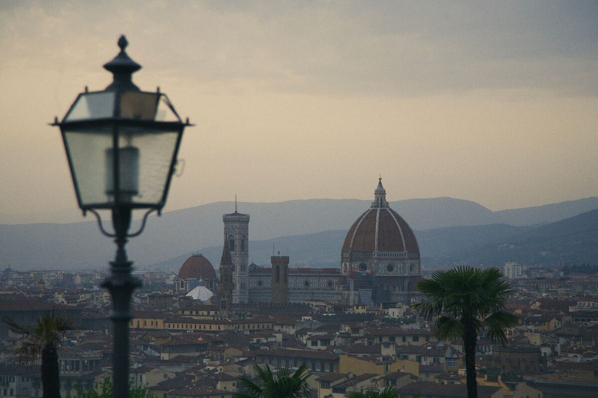 The dome of Florence Cathedral and Giotto's bell tower seen across the city at dusk from Piazzale Michelangelo, framed by an old street lamp and palm trees