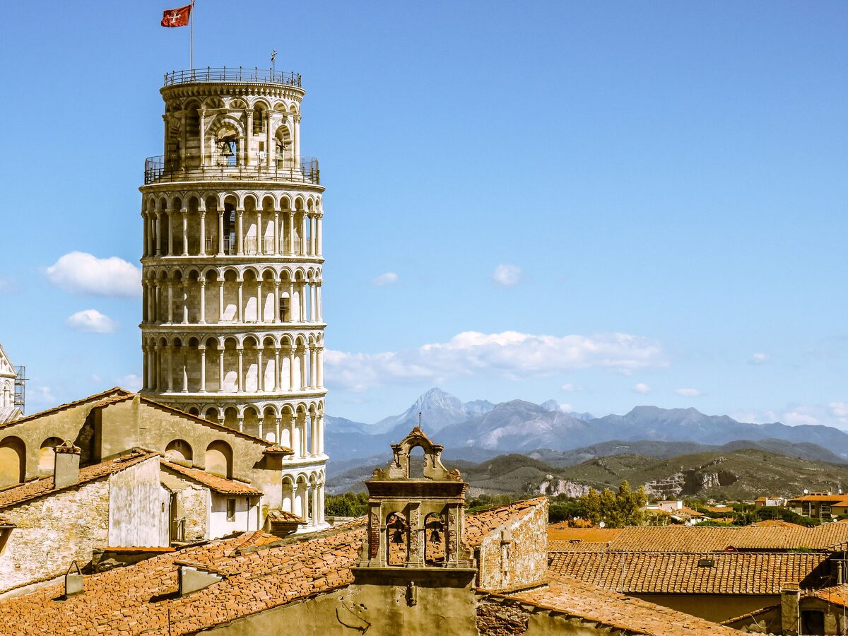 The Leaning Tower of Pisa above old tiled rooftops, with mountains in the distance under a blue sky