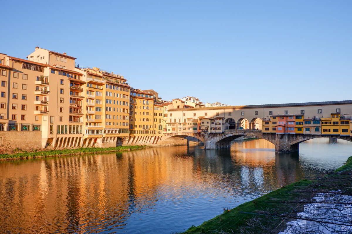 The Ponte Vecchio with its little shops hanging over the arches, reflected in the calm Arno at sunset, with tall riverside buildings on the left