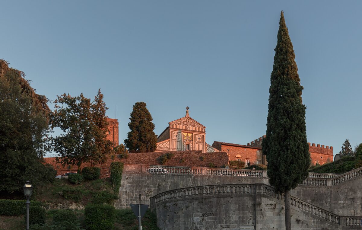 The green and white marble facade of San Miniato al Monte at the top of a hill, with a tall cypress and old walls in front in the evening light