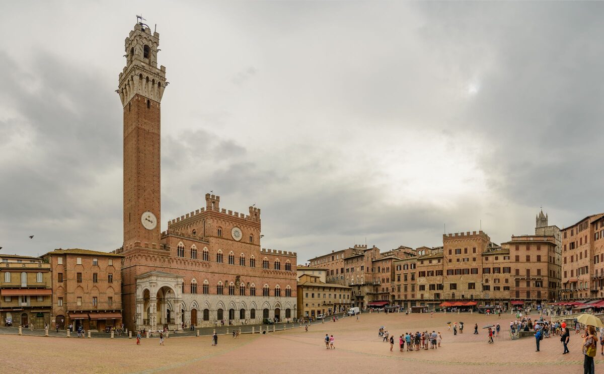 Piazza del Campo in Siena, with the brick Palazzo Pubblico and the tall Torre del Mangia on one side of the sloping square under a cloudy sky
