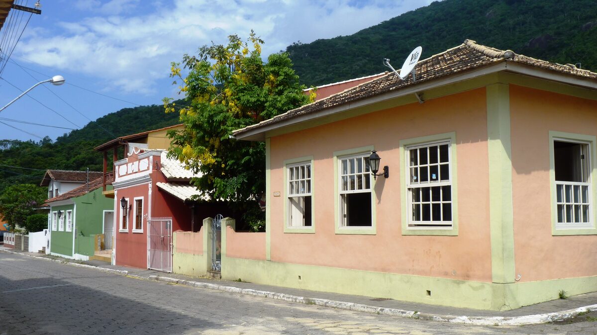 A quiet street of low colonial-style houses in pastel pink, green and red in Caieira da Barra do Sul, with forested hills behind