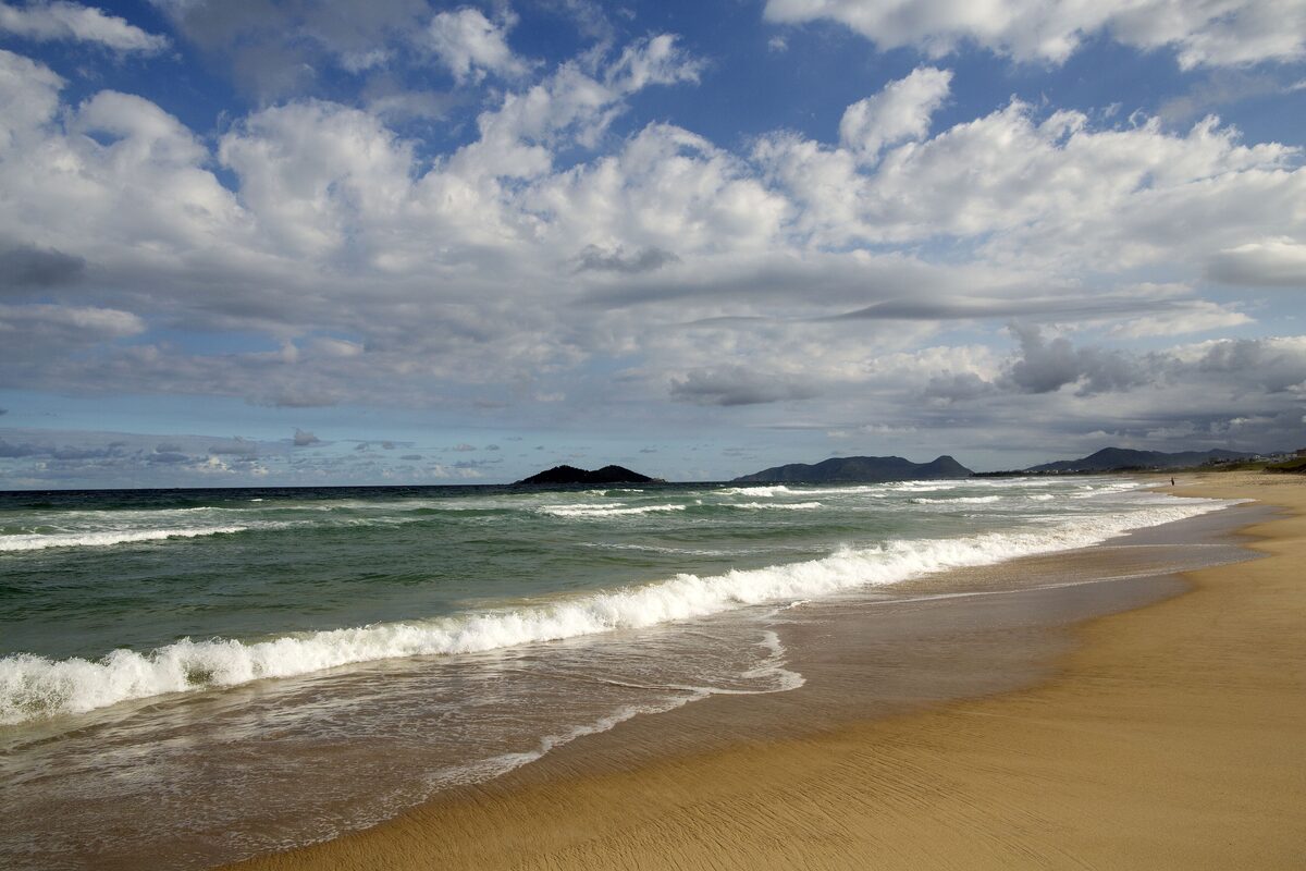 Campeche beach under a blue sky with big white clouds: a long stretch of golden sand, green surf rolling in from the Atlantic and low islands and hills on the horizon