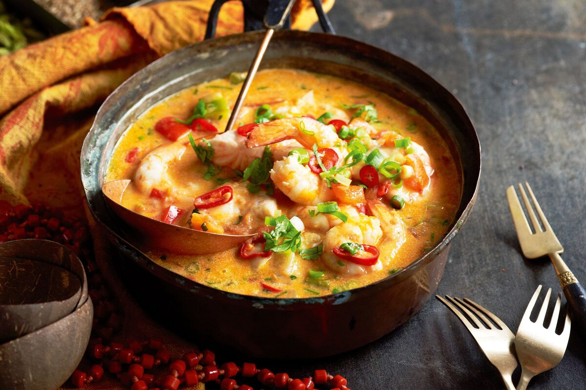A moqueca of shrimp in a creamy orange sauce with chilli and herbs, served in a copper pan beside coconut shells