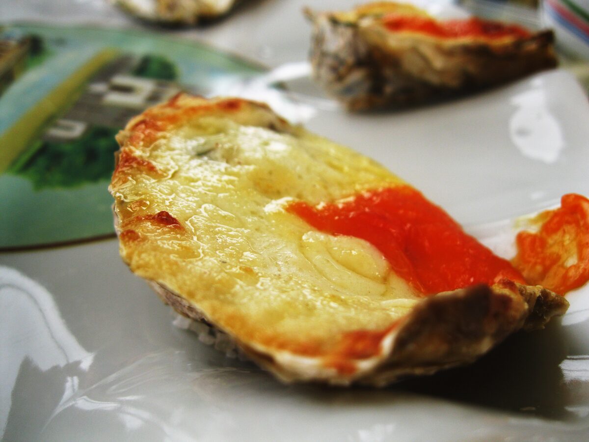 A close-up of a gratinated oyster on the half shell, with melted cheese and a red topping, a dish served in Florianópolis