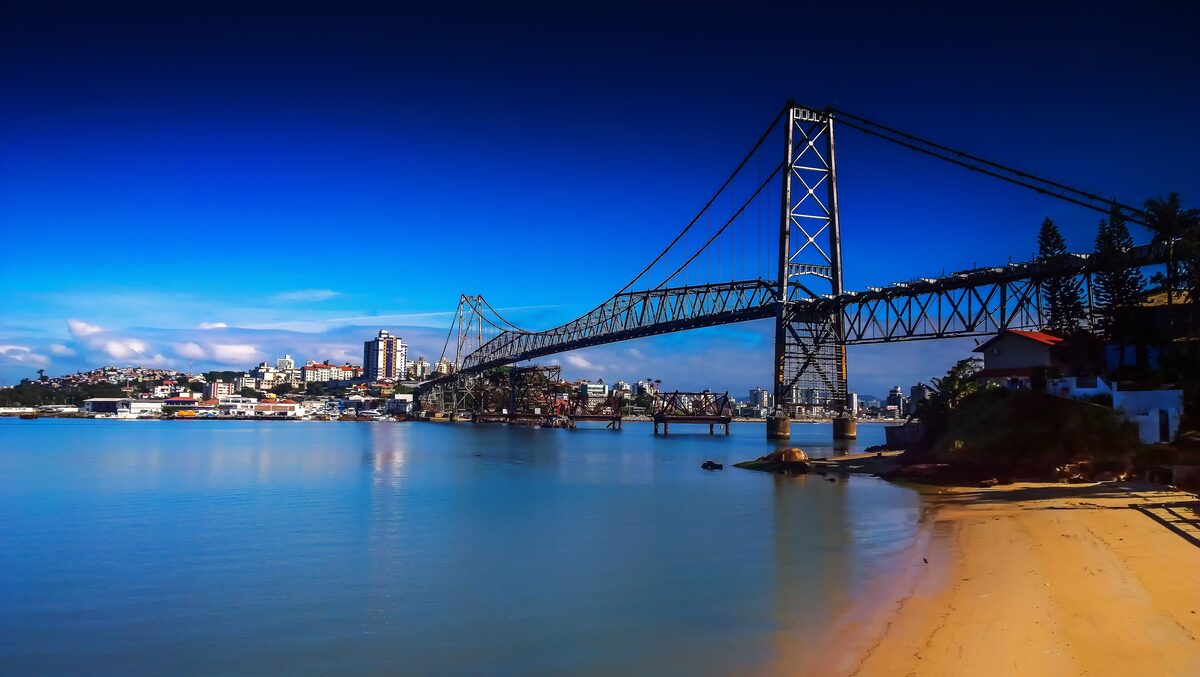 The Hercílio Luz Bridge under a deep blue sky, with the city across the water and sand in the foreground