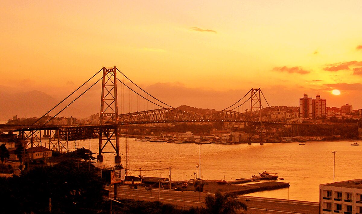 The Hercílio Luz Bridge in silhouette against an orange sunset, with the bay and the city skyline behind