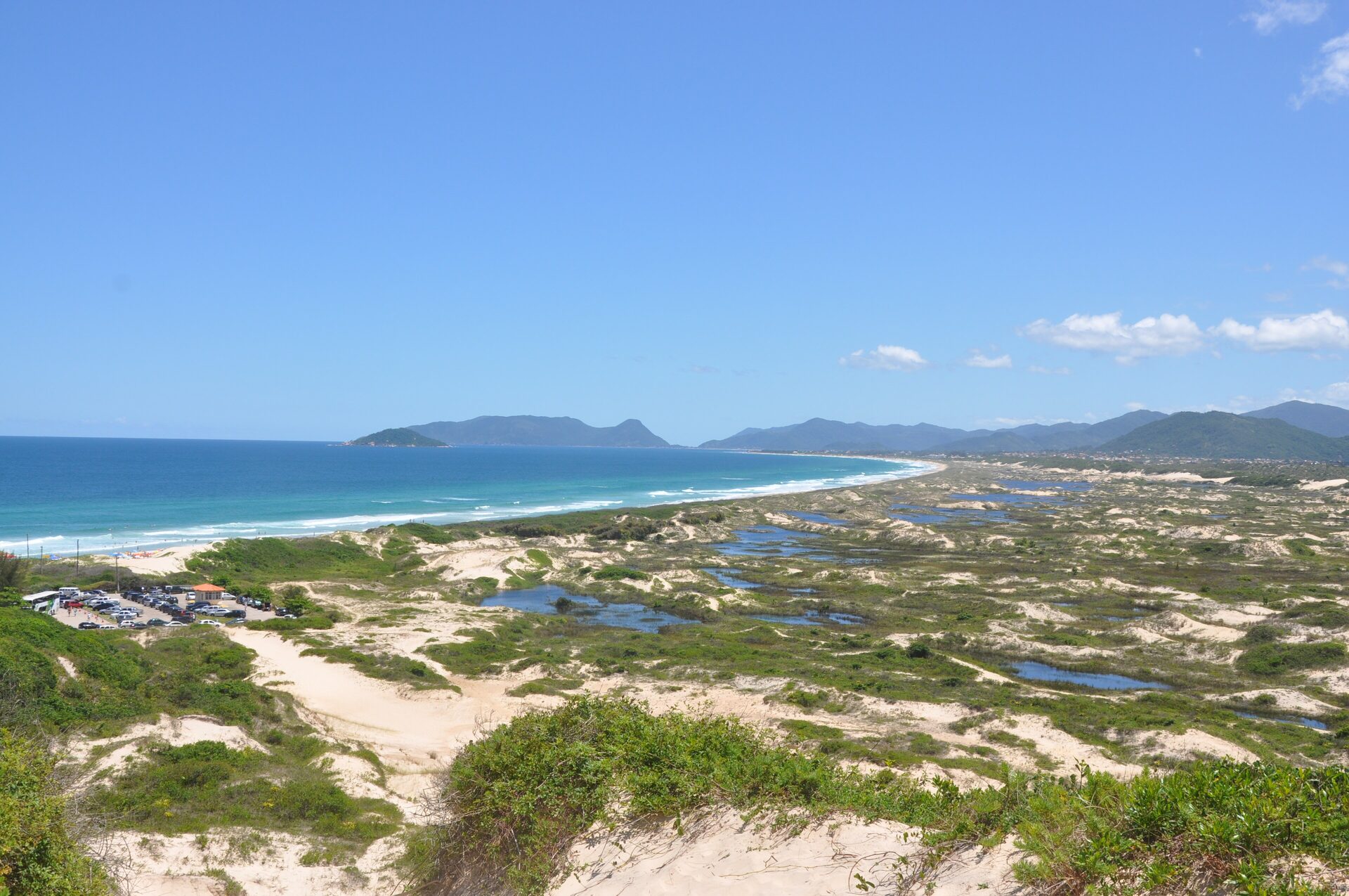 Dune ponds and bare sand above Joaquina beach, with the open Atlantic and islands on the horizon in bright sunshine