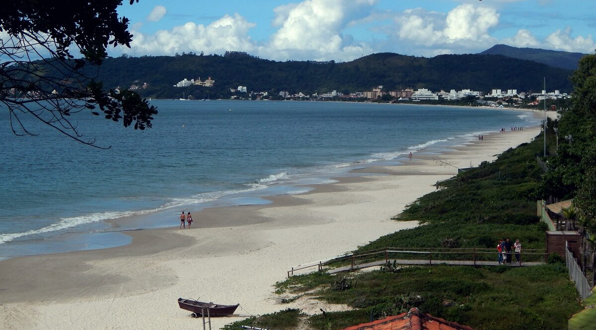 Jurerê beach seen from a raised viewpoint: a long curve of pale sand with a few walkers, calm blue water and the green hills of the bay under a sunny sky