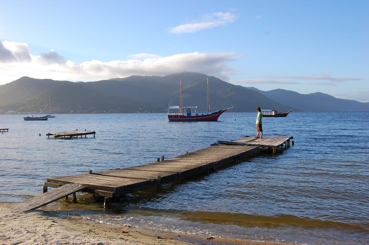 A wooden jetty and a two-masted tourist boat on the Lagoa da Conceição in soft late-afternoon light, with a person at the end of the jetty and hills across the water
