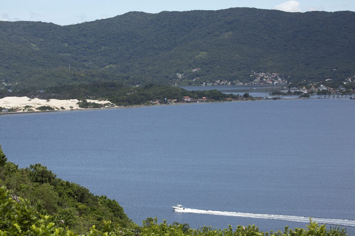 Lagoa da Conceição from a hillside viewpoint: calm blue lagoon water, white dunes on the far shore and a speedboat crossing toward the village