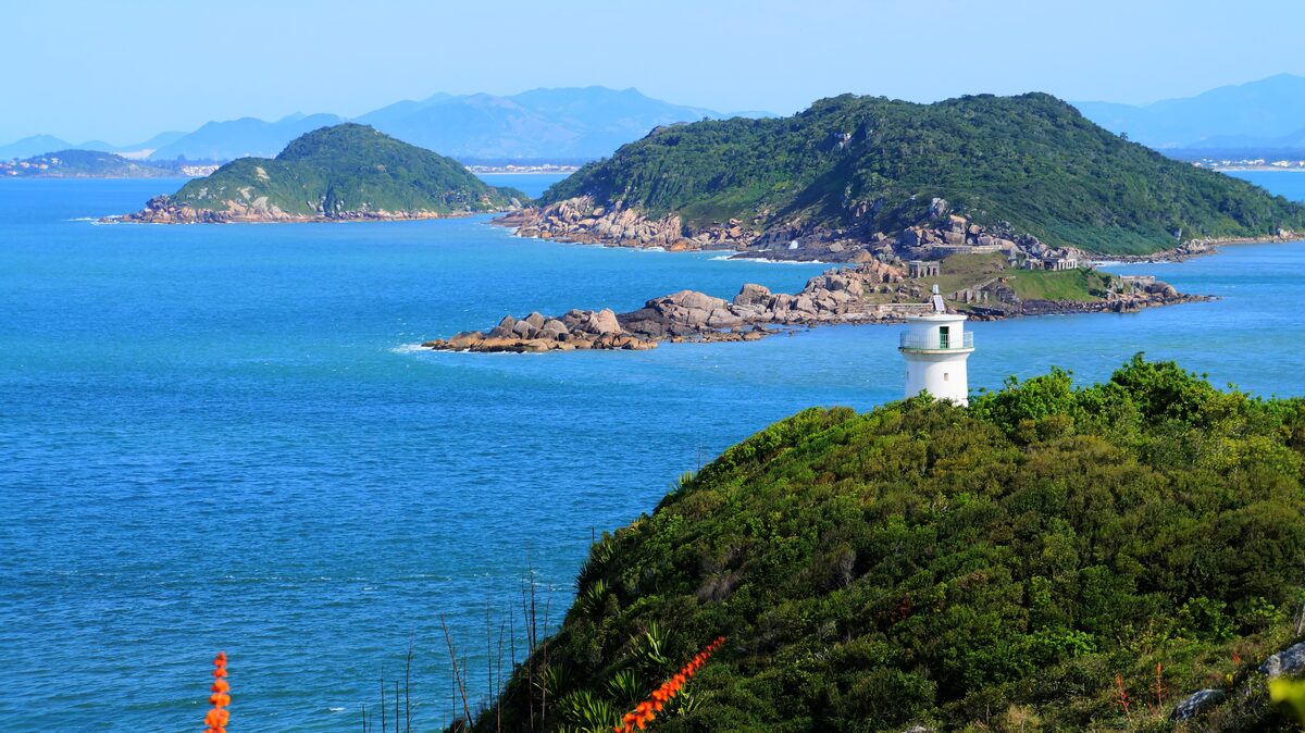 The white Naufragados lighthouse on a green headland above bright blue water, with rocky islets and distant hills