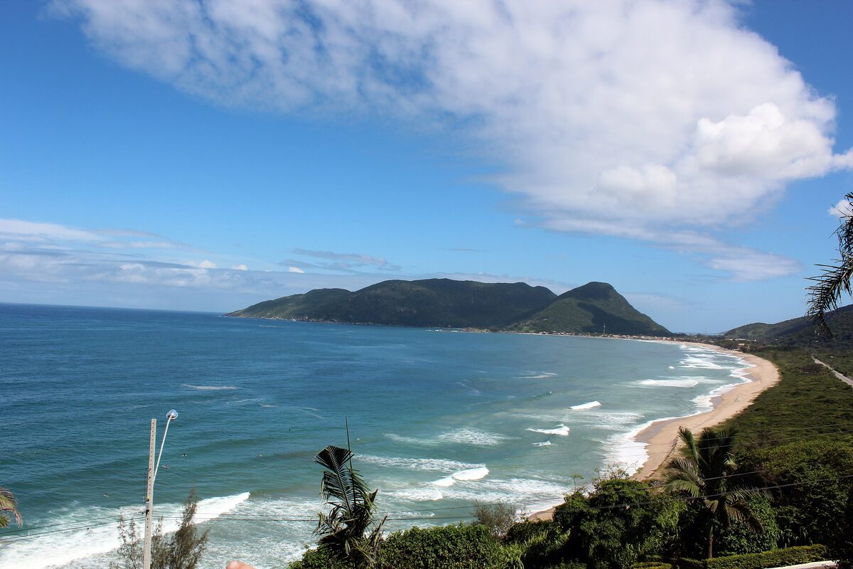 Praia Brava from the hillside: a long curved beach, a green headland and surf in bright daylight