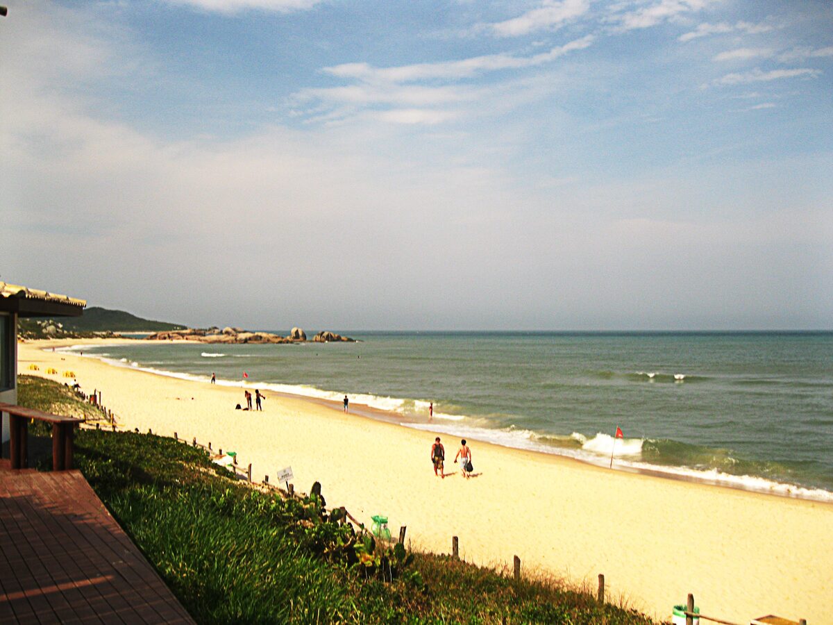 Praia Mole seen from a wooden deck: a wide strip of golden sand with a few walkers, green surf and a rocky headland under a hazy blue sky