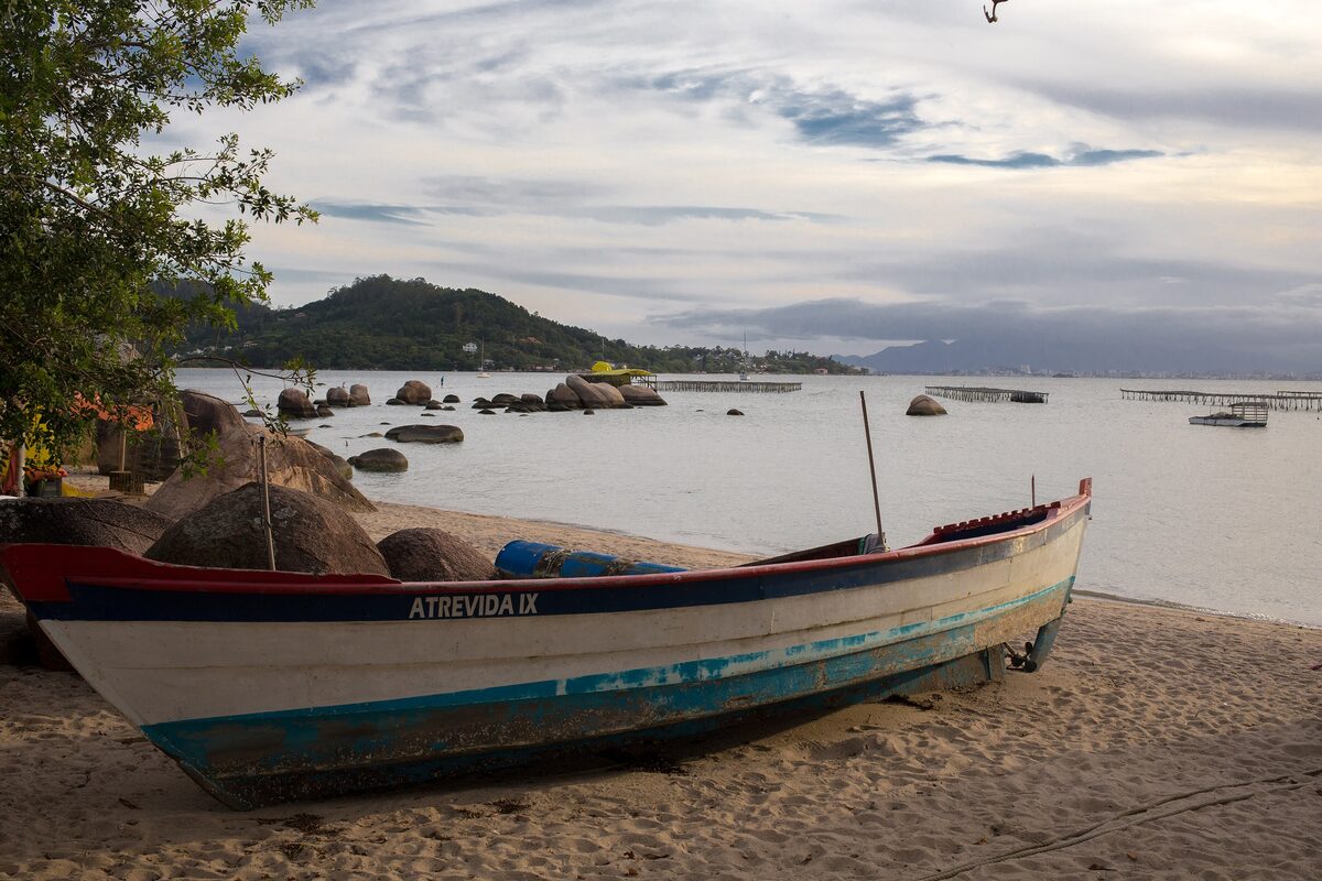 A painted fishing boat pulled up on the sand at Santo Antônio de Lisboa, with rocks and farm racks in the bay behind