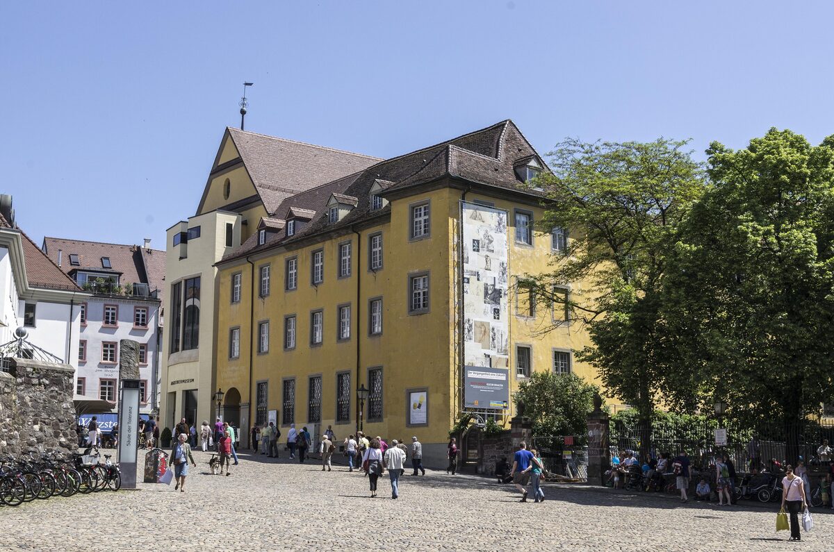 The yellow Augustinermuseum building on Augustinerplatz with people crossing the cobbled square