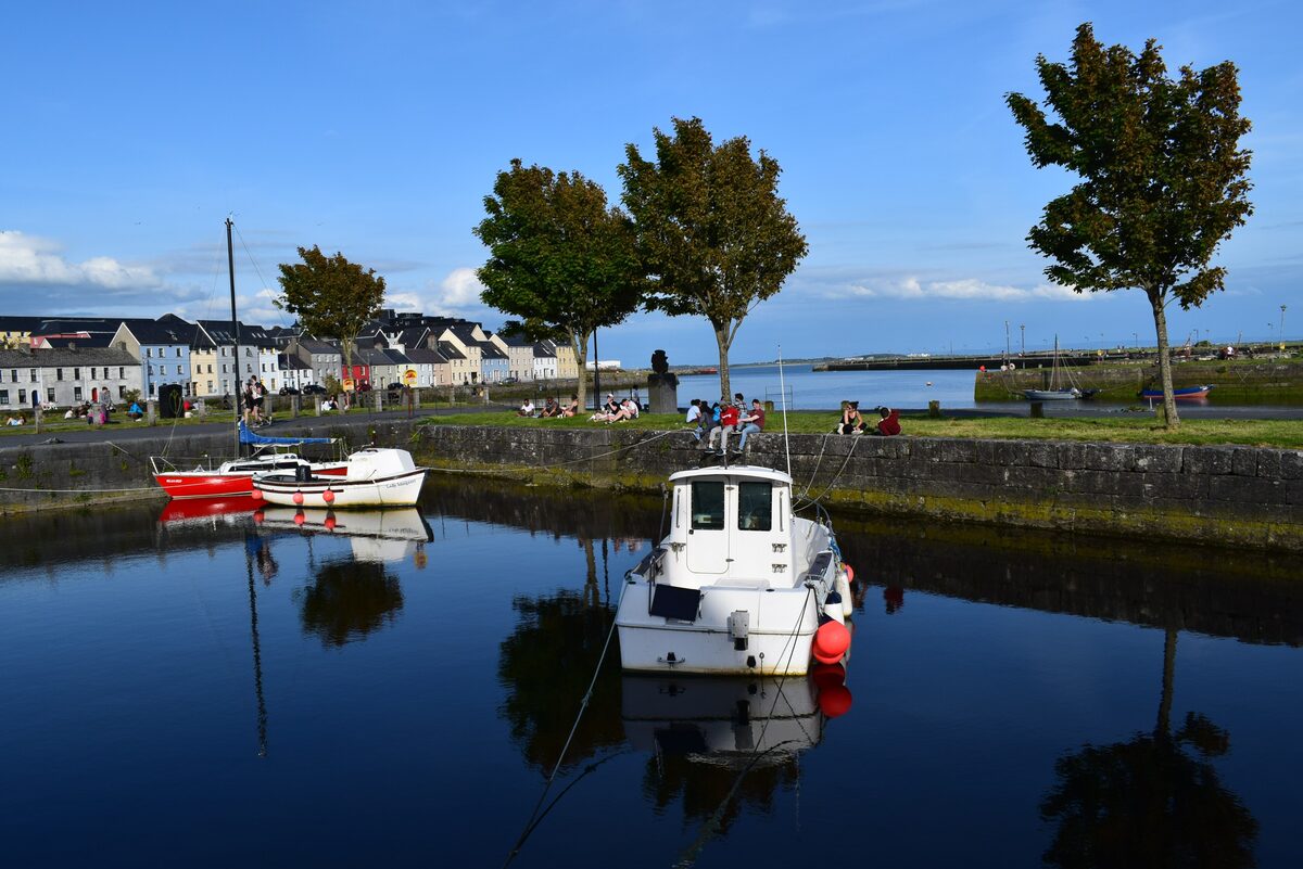 Boats moored in the still water of the Claddagh Basin under trees, with the colored houses of the Long Walk in the background