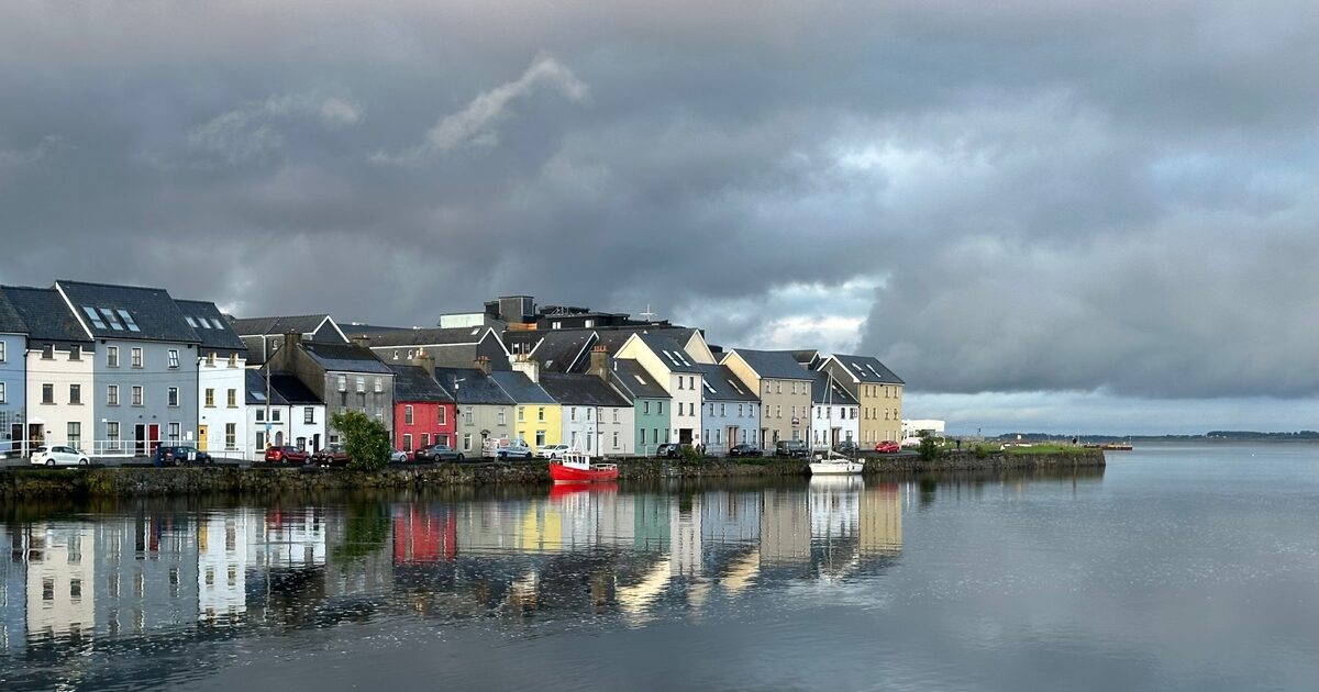 The Long Walk in Galway, a row of colorful painted houses on the quay reflected in calm water under dark gray clouds