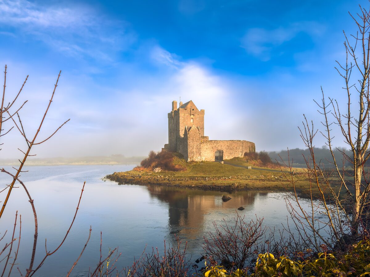 Dunguaire Castle, a stone tower house on a rocky point in Kinvara Bay, reflected in still water under a blue sky