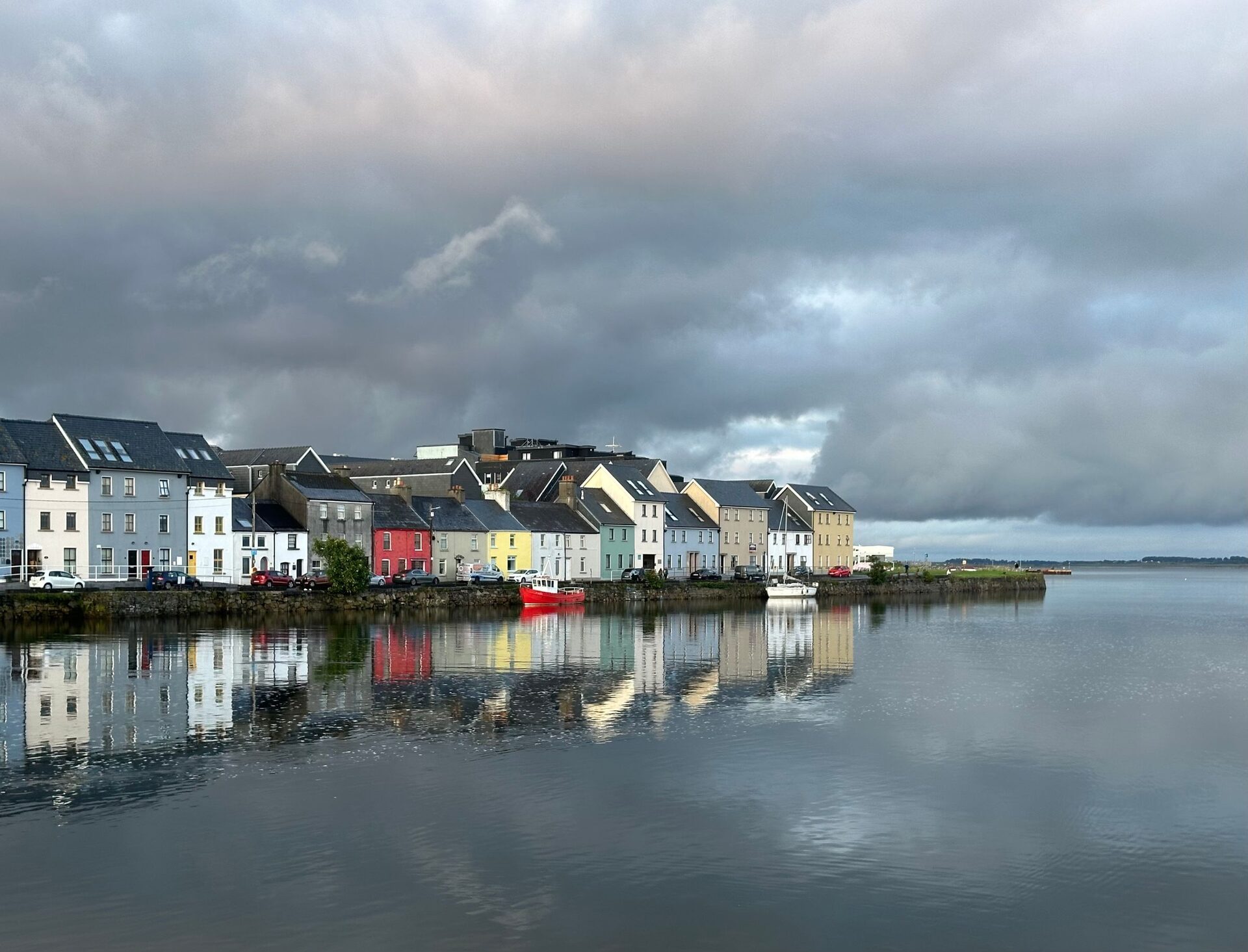 The Long Walk in Galway, a row of colorful painted houses on the quay reflected in calm water under dark gray clouds