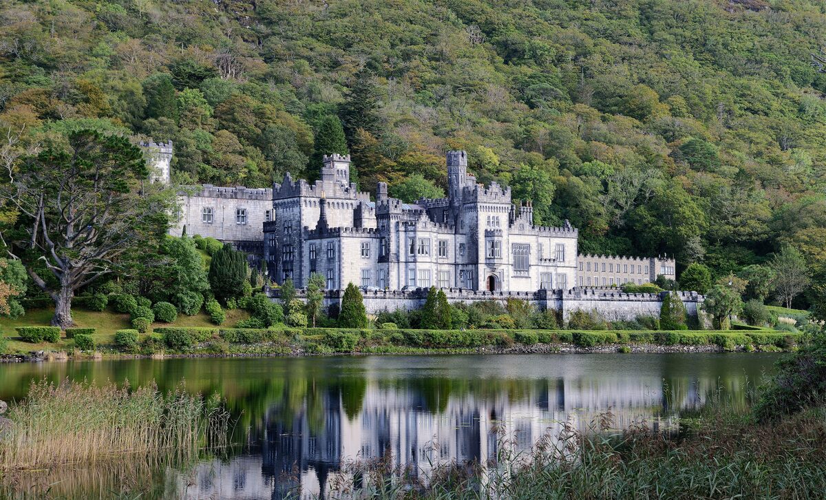 Kylemore Abbey, a pale gray neo-Gothic castle at the foot of a wooded mountain, reflected in a lake