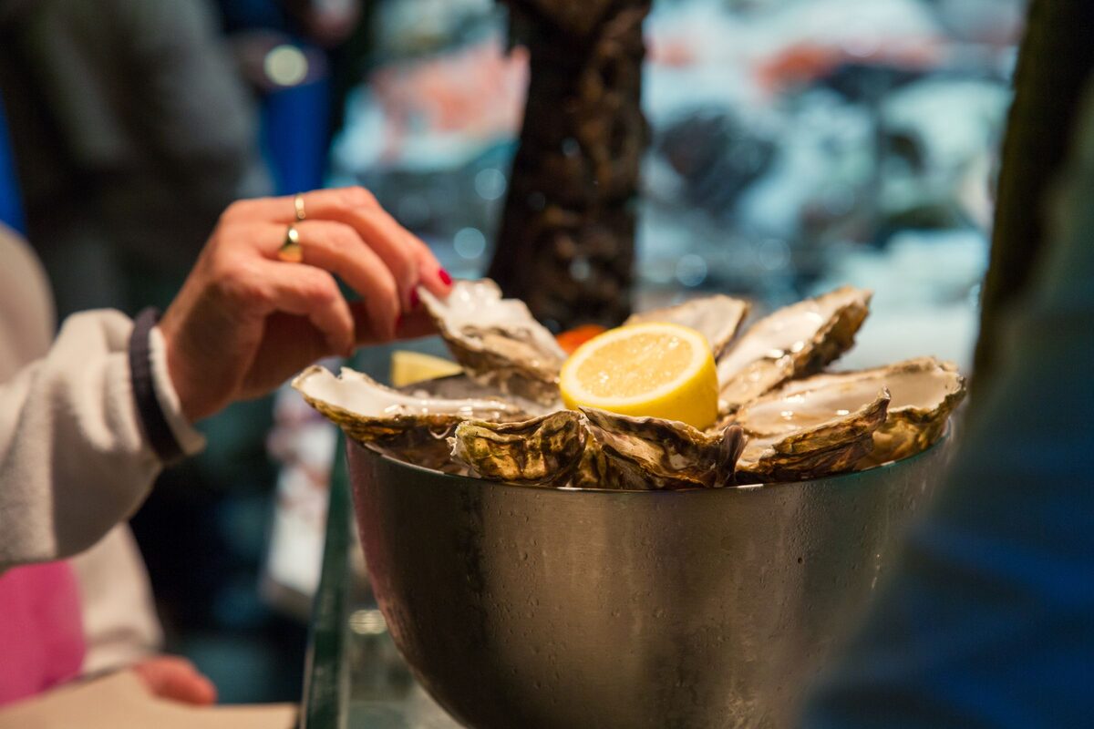 A hand reaching for a fresh oyster from a metal bowl of oysters with a lemon half, in a seafood restaurant