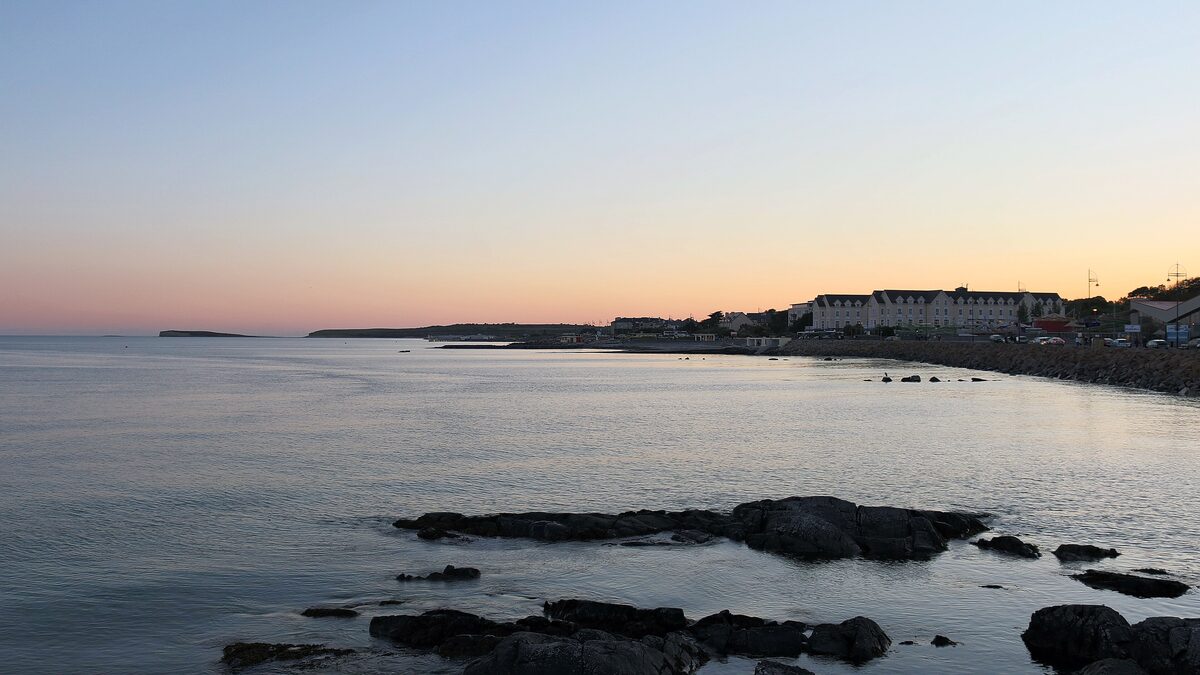 Galway Bay at sunset from the shore at Salthill, with dark rocks in calm water and the lights of the town along the coast