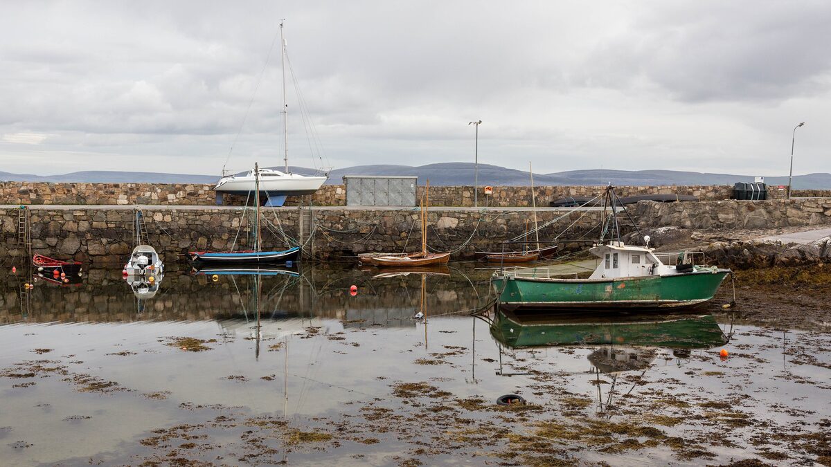 The small stone harbor at Spiddal at low tide, with a green fishing boat and sailing boats, and the hills of the Burren across Galway Bay