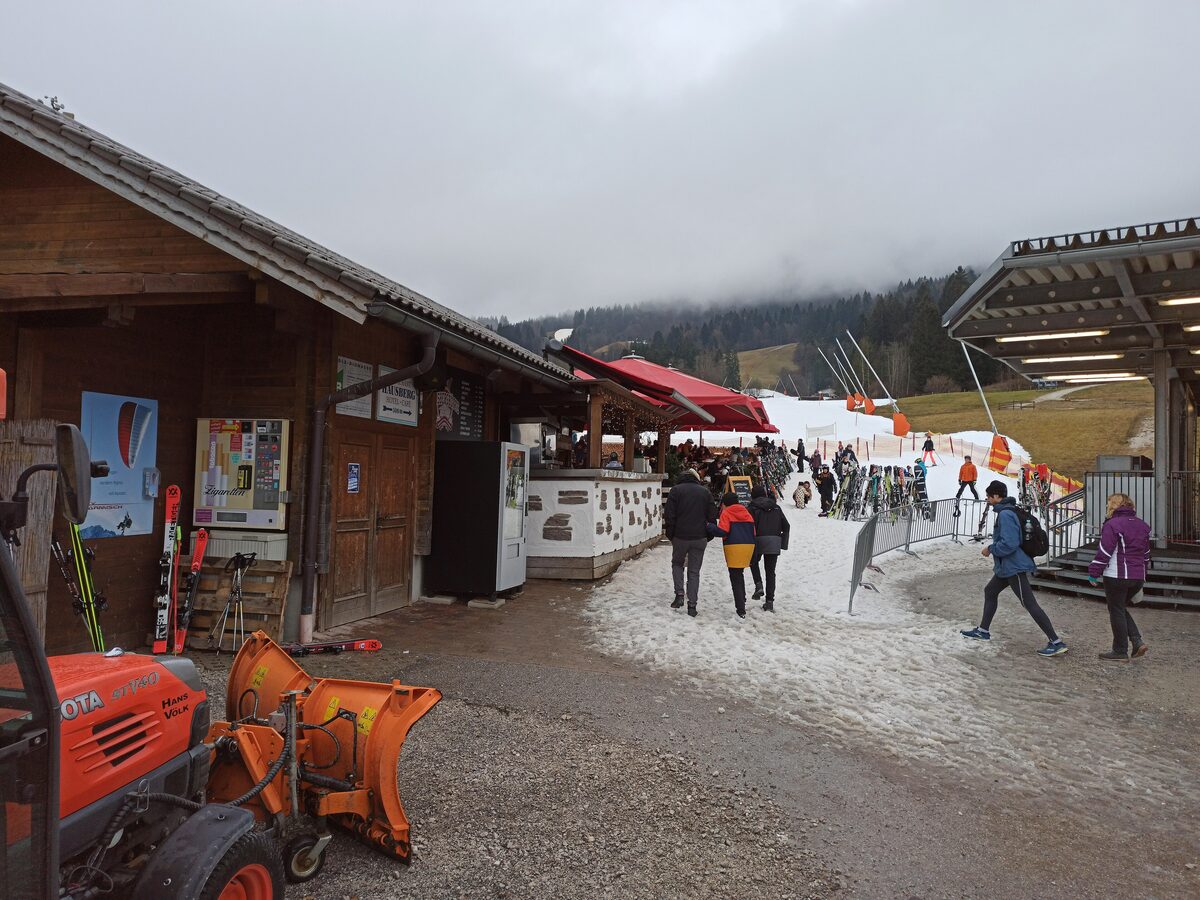 Snow and winter fog at the foot of the Hausbergbahn, with a snow plough and skiers walking to the lift