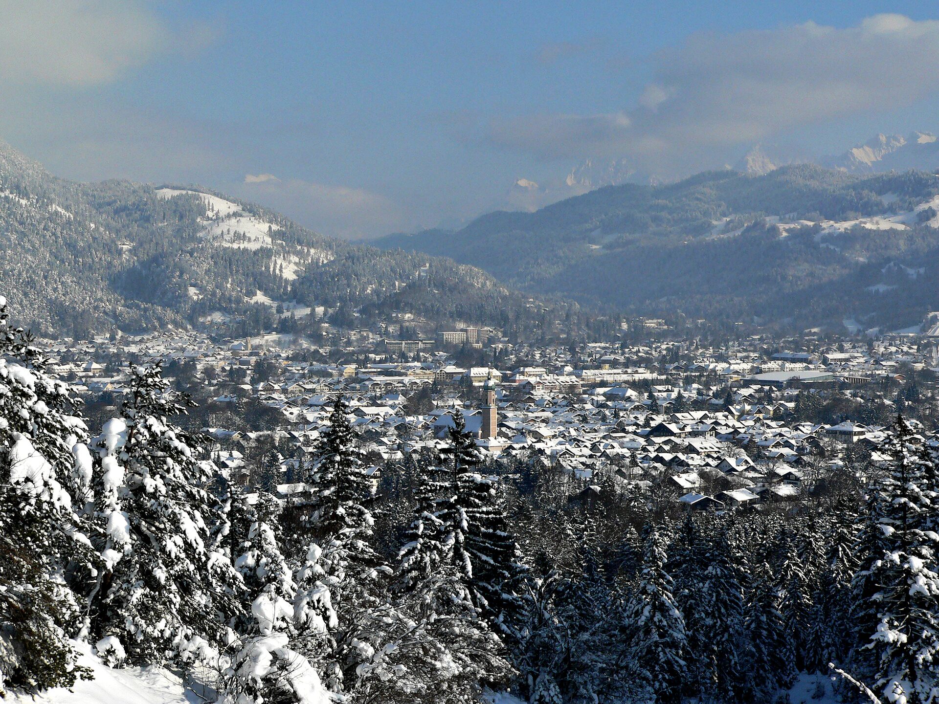 Snowy rooftops of Garmisch-Partenkirchen seen through snowy firs, with the Wetterstein ridge hazy behind