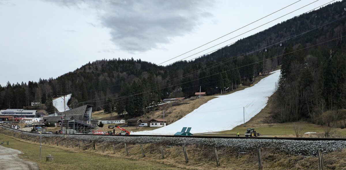 A single strip of machine snow on the Kreuzeck slope above the valley in February, with the railway and grooming vehicles at its foot