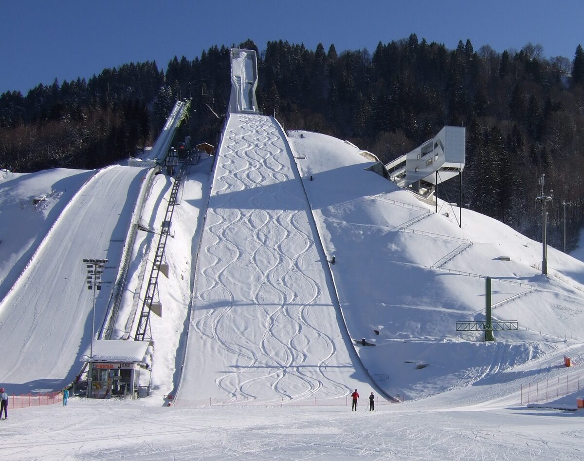 The Große Olympiaschanze covered in snow under a blue sky, with the jump tower above the landing slope and two people standing at the bottom