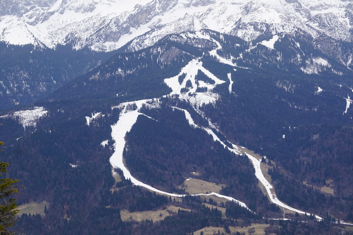 Thin snow strips of the Hausberg ski runs winding through dark forest, seen from above, with snowy Wetterstein peaks at the top