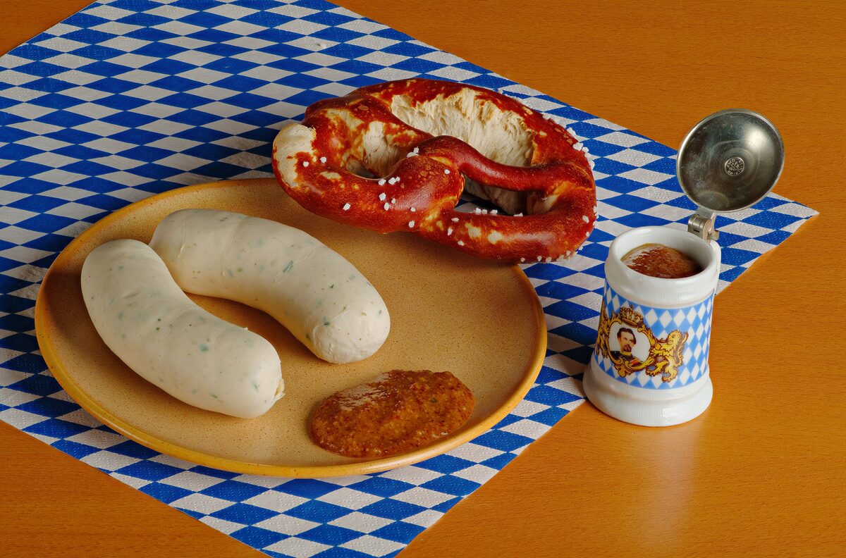Two white sausages on a plate with a pretzel and sweet mustard, on a blue-and-white checked cloth